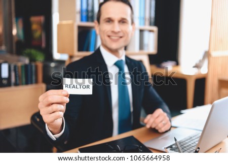 Realtor in suit sitting at desk in office. Man is posing on camera with realtor sign.