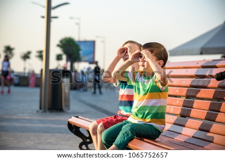 Toddlers boy and his sibling brother sitting on a bench by the city and look for something interesting. Summer time hot evening, hugging each other, friendship concept.