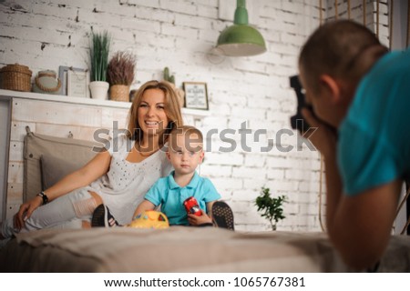 Father taking a photo of his smiley wife and little son, who sitting on the bed