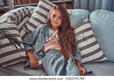 Portrait of a cute little girl with long brown hair and piercing glance, looking at a camera, lying on a sofa at home alone