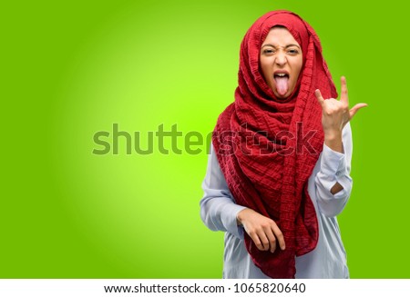 Young arab woman wearing hijab making rock symbol with hands, shouting and celebrating with tongue out