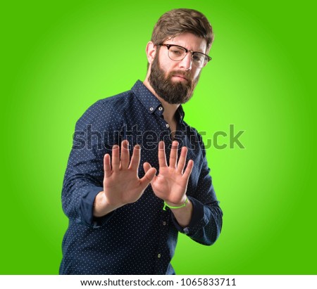 Young hipster man with big beard annoyed with bad attitude making stop sign with hand, saying no, expressing security, defense or restriction, maybe pushing over green background