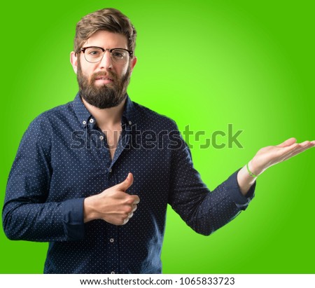 Young hipster man with big beard thumb up holding something in his empty hand and making ok gesture over green background