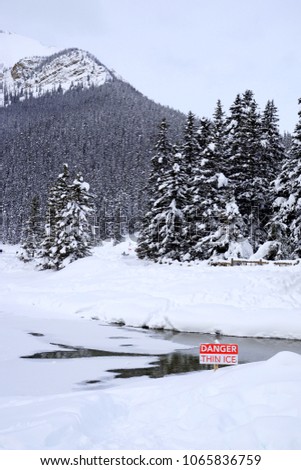 A sign warns of thin ice on a lake with snow covered trees and mountains in the background