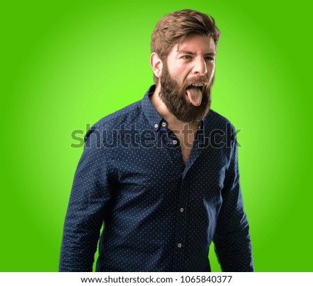 Young hipster man with big beard sticking out tongue at camera at sign of disobedience, protest and disrespect over green background