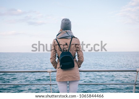 Back view of young woman wearing beige coat, scarf, hat and backpack looking at sea. Female traveller holds her hands in pockets stands at quay.