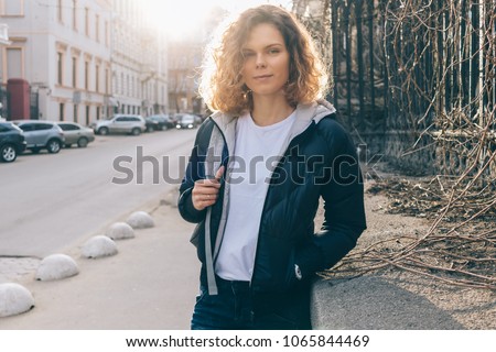 Young woman traveler looks at camera, smiles while standing on old town street. Candid image of girl wearing jacket, white t-shirt and jeans holds backpack strap, morning sun shines through her hair.