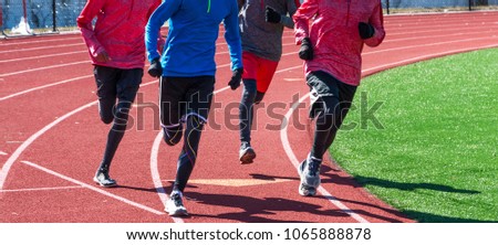 High school boys run a workout together wearing gloves and spandex on a red track during winter track and field practice. Royalty-Free Stock Photo #1065888878