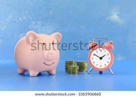 Piggy bank on a saturated blue background. Nearby is a small pink alarm clock and there are three stacks of coins between them. Beautiful bright photo.