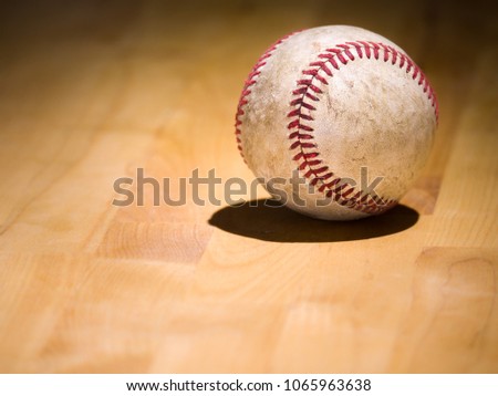Close up sports background image of an old used weathered leather baseball showing intricate detailing and red laces sitting on a wood butcher block counter with lighting from behind and dark shadows.