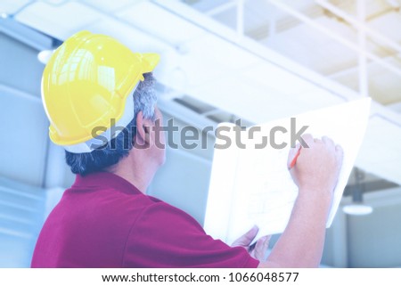 asian engineer in yellow hard safety helmet is checking the structure of building and factory blueprint on the factory construction site with sunbeam. 