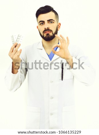 Doctor with beard holds pills blisters and shows ok sign. Treatment and ambulance services concept. Man with serious face in white hospital gown. Physician with drugs isolated on white background