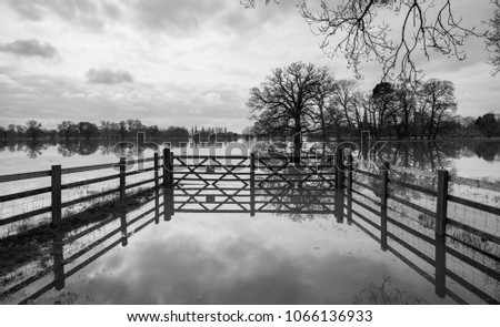 black and white picture of flooded park in the UK with trees partially submerged under water and nice reflections of fence and gate with stormy skies in background