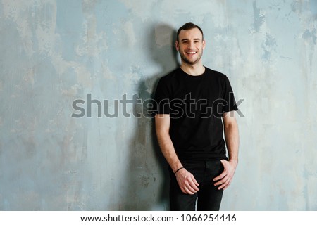 portrait of a young man in a black T-shirt leaned against a vintage gray wall. Copy space. Masculine model of bristles. Happy smiling. Royalty-Free Stock Photo #1066254446