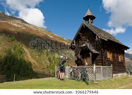 man with electric bicycle, ebike, take a photo of old wooden church of small hut village, cabin, Valais, snow covered mountains, autumn colors, sun, clouds, travel, above city of Goppenstein, Swiss
