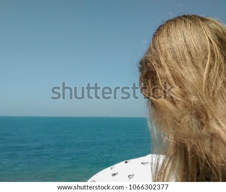 View from behind: head of blond girl in white shirt with black hearts, looking at the ocean - hair blowing in the wind, with blue water and sky filling much of the picture, leaving space to add text
