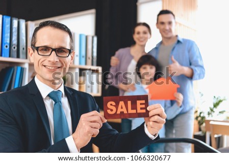 Realtor in suit sitting at desk in office. Realtor is presenting sale sign with family in background.