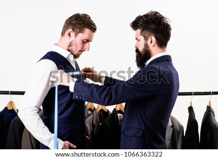 Businessman, client stand near hanger with suits while tailor working. Tailor measure chest for sewing suit, white background. Tailors work concept. Man with measuring tape taking measurements.