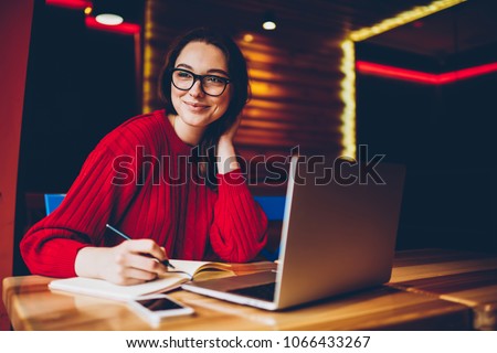 Smiling young woman enjoying freelance work in cafe interior making notes of ideas in notepad, positive female student doing homework in good mood satisfied with wifi for learning on laptop computer Royalty-Free Stock Photo #1066433267