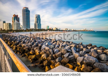 Long exposure of Miami Beach pier, mole and buildings with bathing activity on the beach and blurred clouds
