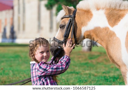 Beautiful little blonde girl embraces horse pony. She has happy fun smiling face, pretty blue eyes, plaid shirt. Child and animals portrait. Creative amazing concept. Happy couple friends.