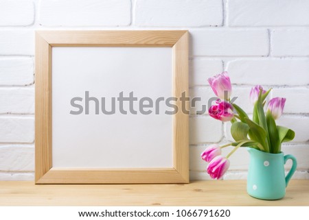 Wooden square picture frame mockup with pink tulip in mint pitcher vase near painted brick wall. Empty frame mock up for presentation design. Template framing for modern art.
