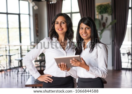Two cheerful female colleagues using tablet computer in cafe. They are standing and looking at camera with cafe interior and windows in background. Technology concept. Front view.