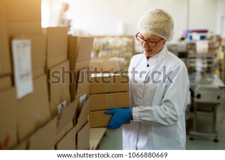 Young female cheerful worker is picking up a cardboard box stack from a bigger stack in a factory production line. Royalty-Free Stock Photo #1066880669
