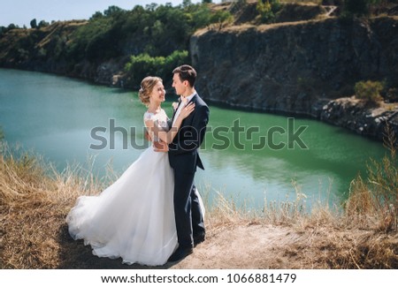 Very gentle and smiling bride and groom in a lace dress, embracing against the background of rocks, green trees and a green lake. Sunny wedding day.