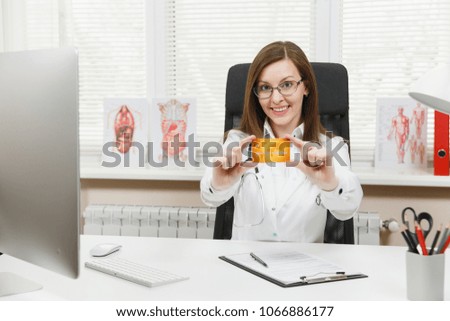 Young smiling female doctor sitting at desk, holding credit card, working with medical documents in light office in hospital. Woman in medical gown in consulting room. Healthcare, medicine concept