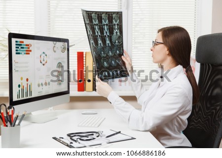 Female doctor sitting at desk with computer, film x-ray the brain by radiographic image ct scan mri in light office in hospital. Woman in medical gown in consulting room. Healthcare, medicine concept
