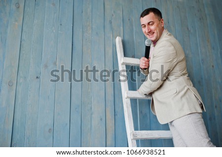 Handsome man in beige suit and pink shirt with microphone against ladder background on studio. Funny face of toastmaster and showman.