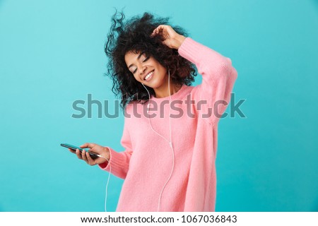 Joyous american woman in casual clothing dancing and listening to music with pleasure via white earphones isolated over blue background Royalty-Free Stock Photo #1067036843