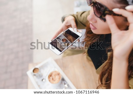 Close-up portrait of concentrated woman with white manicure making picture of her lunch. Overhead photo of girl in sunglasses holding smartphone above coffee and croissant.