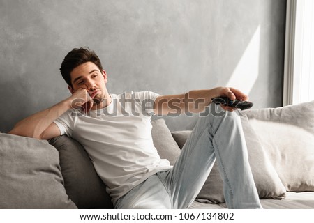 Photo of young bored man sitting on couch at home and changing TV channels with disinterest