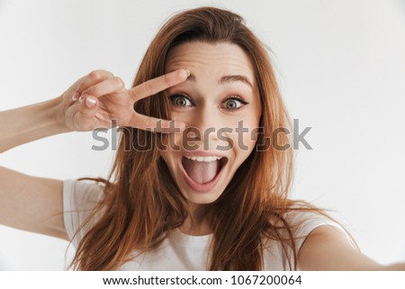 Joyful woman in casual clothes making selfie and showing peace gesture while looking at the camera over grey background