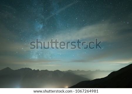 Milky Way galaxy and starry sky from high elevation in summertime on the Alps. Fog and mist, city lights.