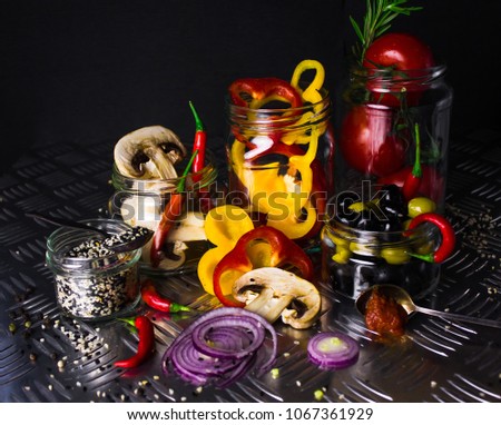 Fresh food ingredients vegetables (tomatoes, bell peppers, mushrooms, olives, onions) for pizza in glass jars dark background