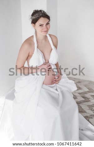 photo Pregnant girl in a diadem in a white drapery on a white background sits
