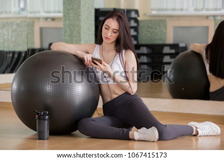 Happy sporty young beautiful girl with smartphone listening to music while sitting near with fitness ball