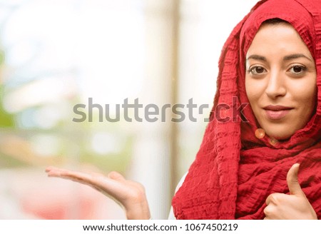 Young arab woman wearing hijab thumb up holding something in his empty hand and making ok gesture