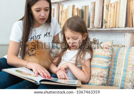 A pretty little girl with brown hair and her mother are reading a book and watching cartoons on computer with their dog. Family free time. Friendship. Pomeranian Spitz.
