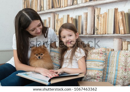 A pretty little girl with brown hair and her mother are reading a book and watching cartoons on computer with their dog. Family free time. Friendship. Pomeranian Spitz.