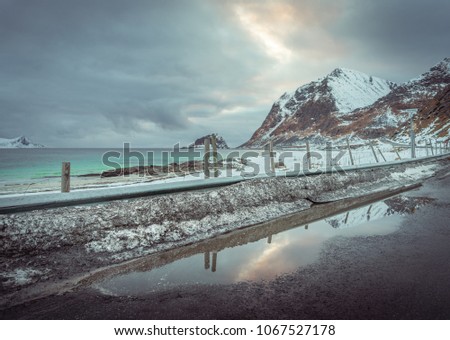 Beach with green and blue sea, contrasty clouds, and mountains in background. road with dirty snow
