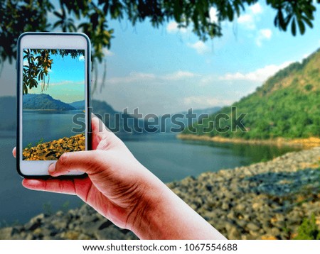 Hand holding smart phone and take a photo of white clouds,river,mountain,blue sky and landscape with blurred natural background | copy space