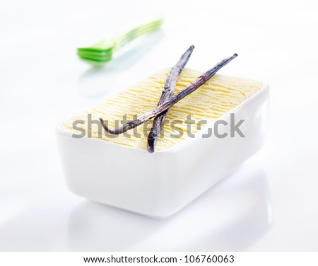 Open tub of creamy vanilla icream with two dried vanilla seedpods used as flavouring in its manufacture on a white background with reflection