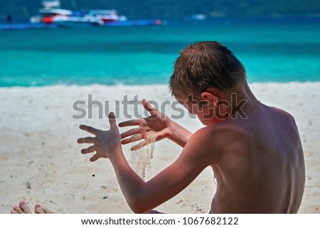 Boy playing with sand on tropical sea beach, run dry sand through your fingers. Rear view against the turquoise sea. Copy space.