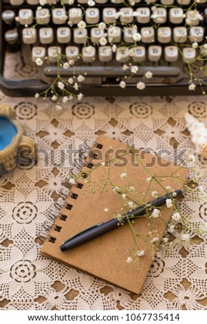 Spring or summer freelance, business or writing concept. Notepad with black pen on lace table cover with retro typewriter blurred at distant view.  Vertical composition.
