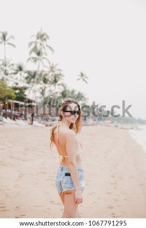 attractive girl standing on sandy beach in bikini top and sunglasses