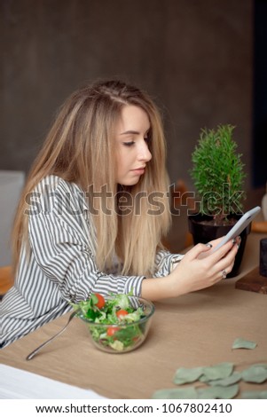 Young woman sitting at the table looks at phone. The woman is going to eat Fresh salad that is on the table in front of her.